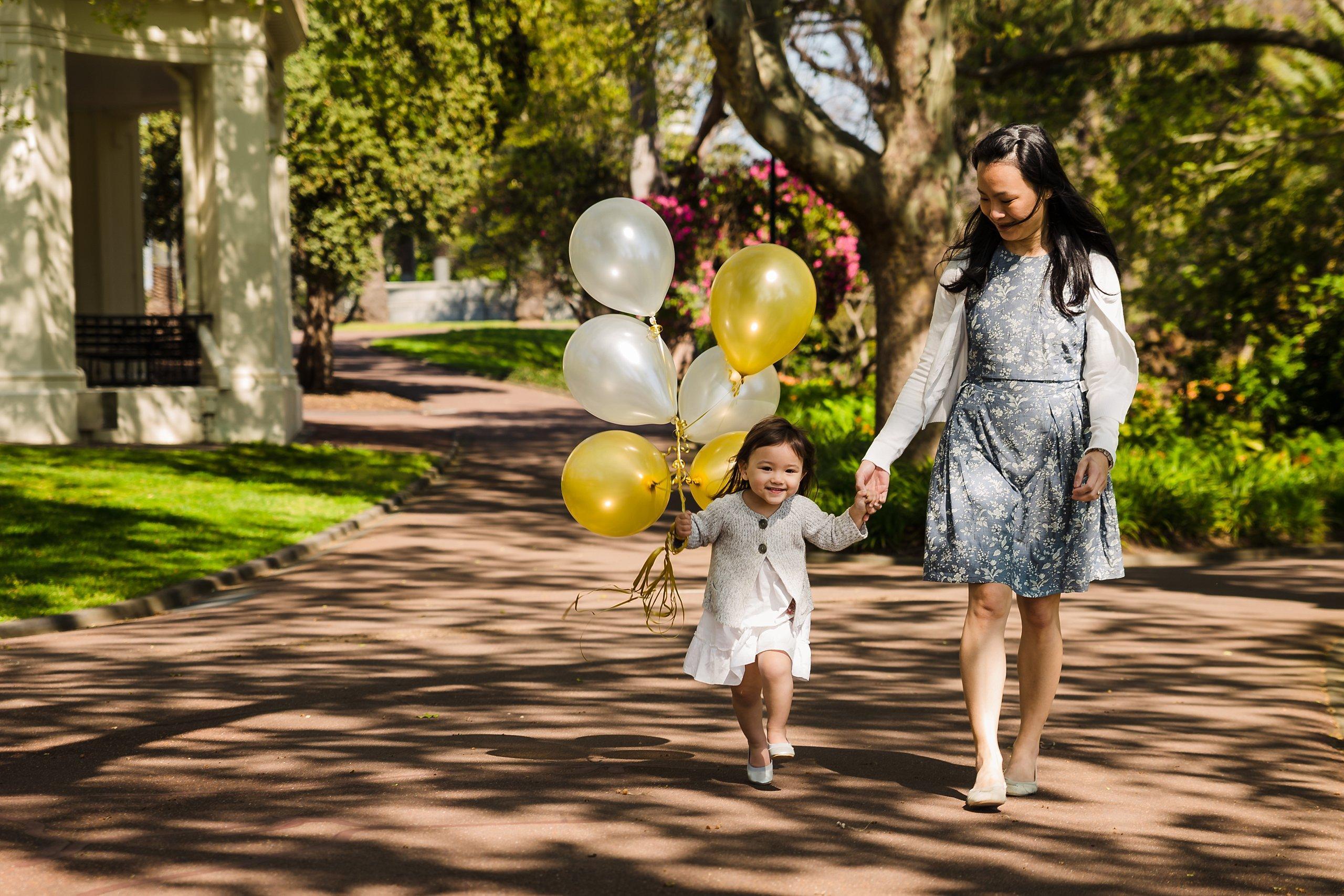 mother holding hands with her younge daughter with yellow and white ballons walking in queen victoria gardens