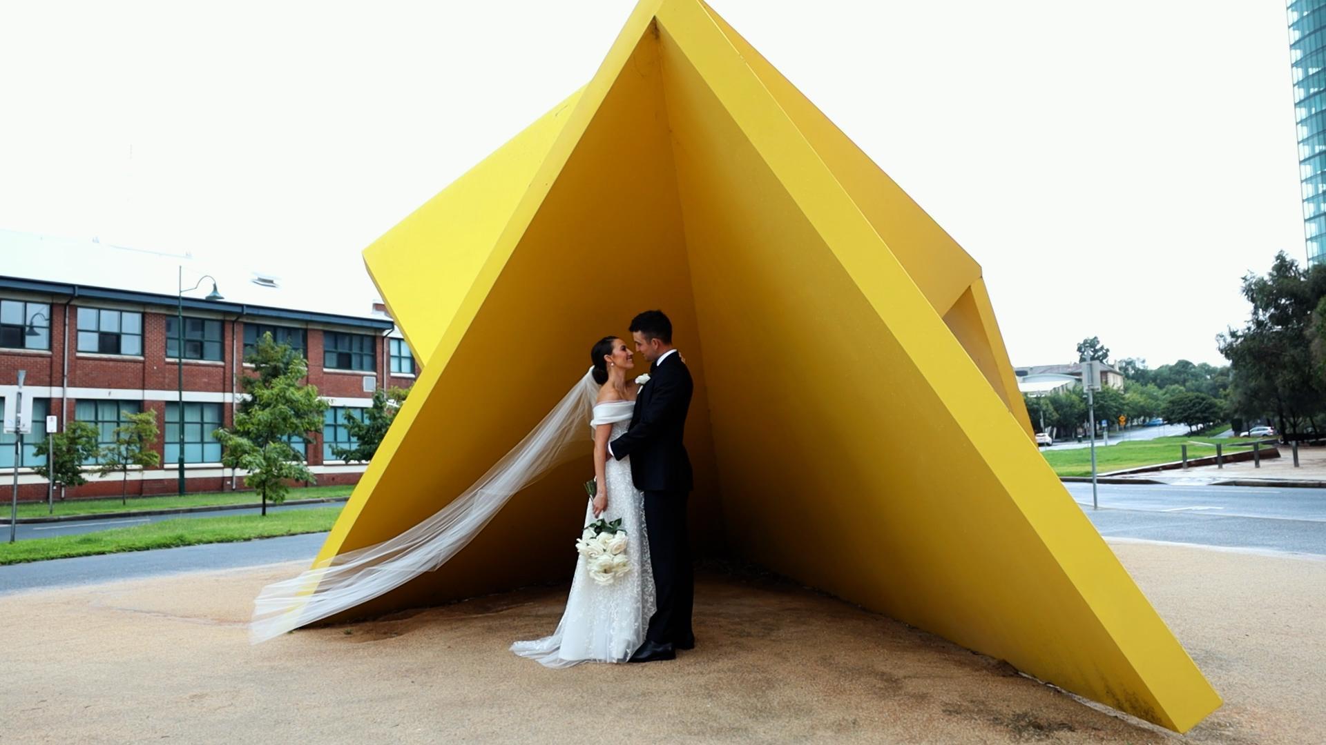 bride and groom smiling at each other under a yellow art sculpture in melbourne
