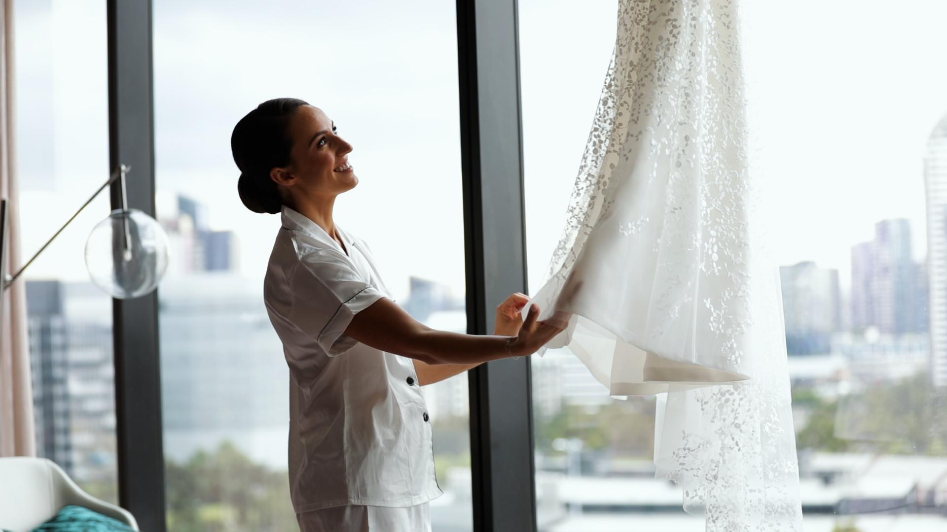 bride smiles while touching her wedding dress hanging up near window in hotel room