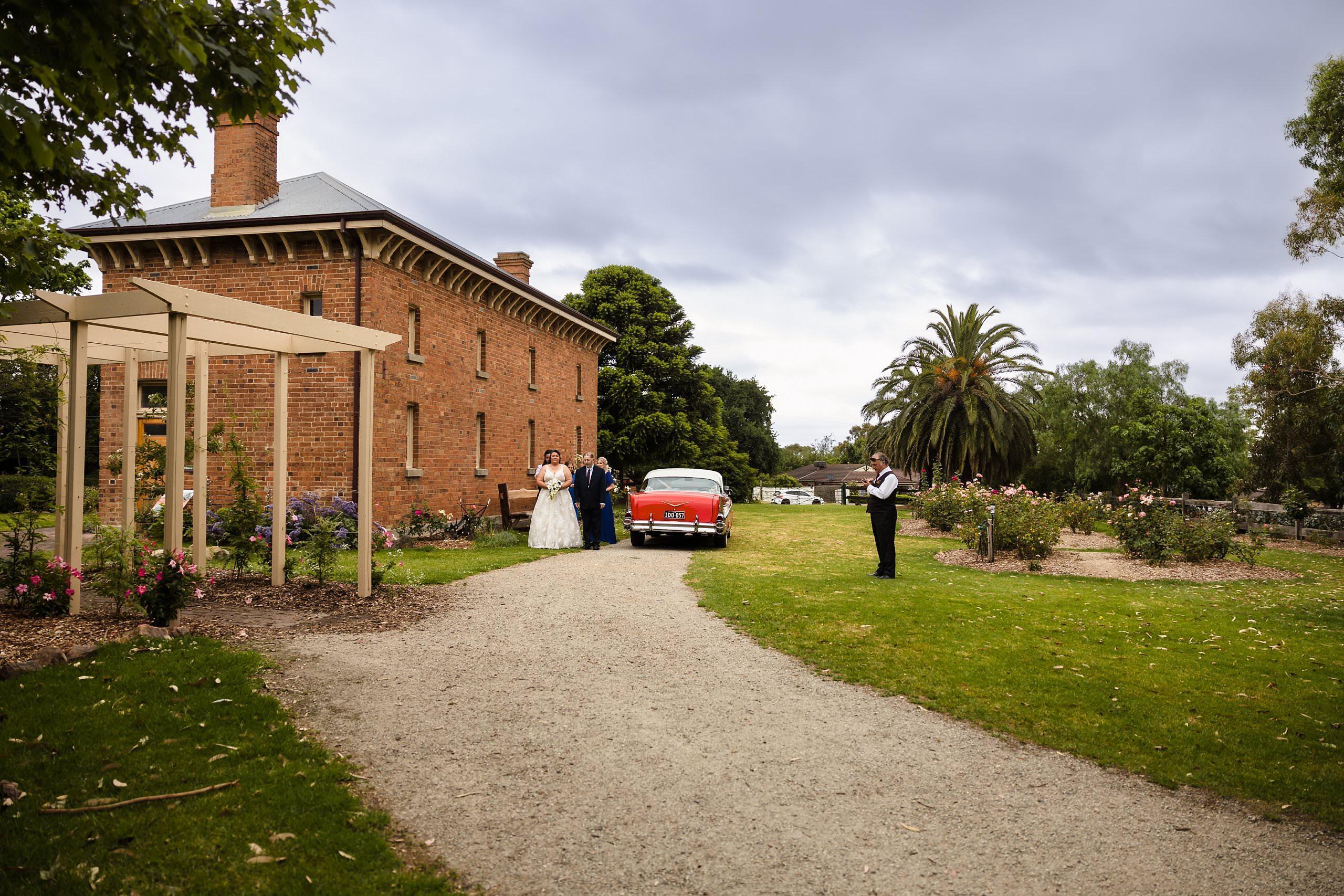 wide view of bride arriving at old cheese factory wedding in a red vintage car