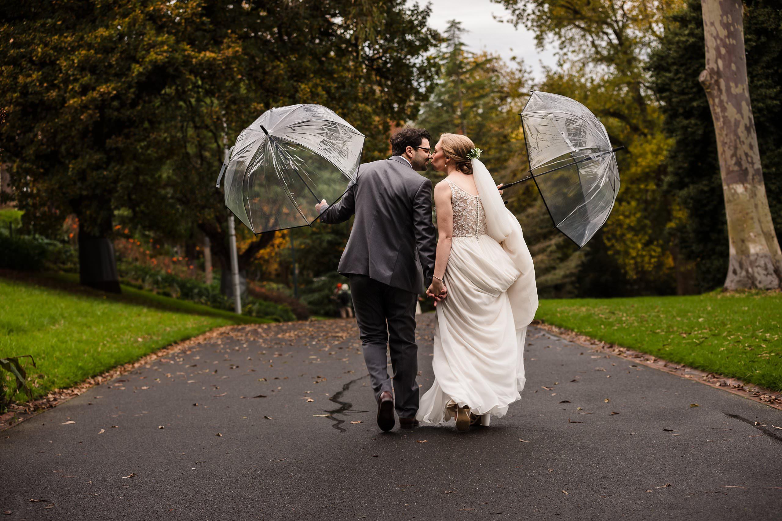 bride and groom holding umbrellas while walking in the treasury gardens during romantic elopement